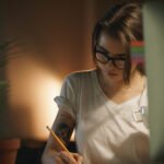 Picture of serious young woman designer sitting indoors at night writing notes using computer. Looking aside.
